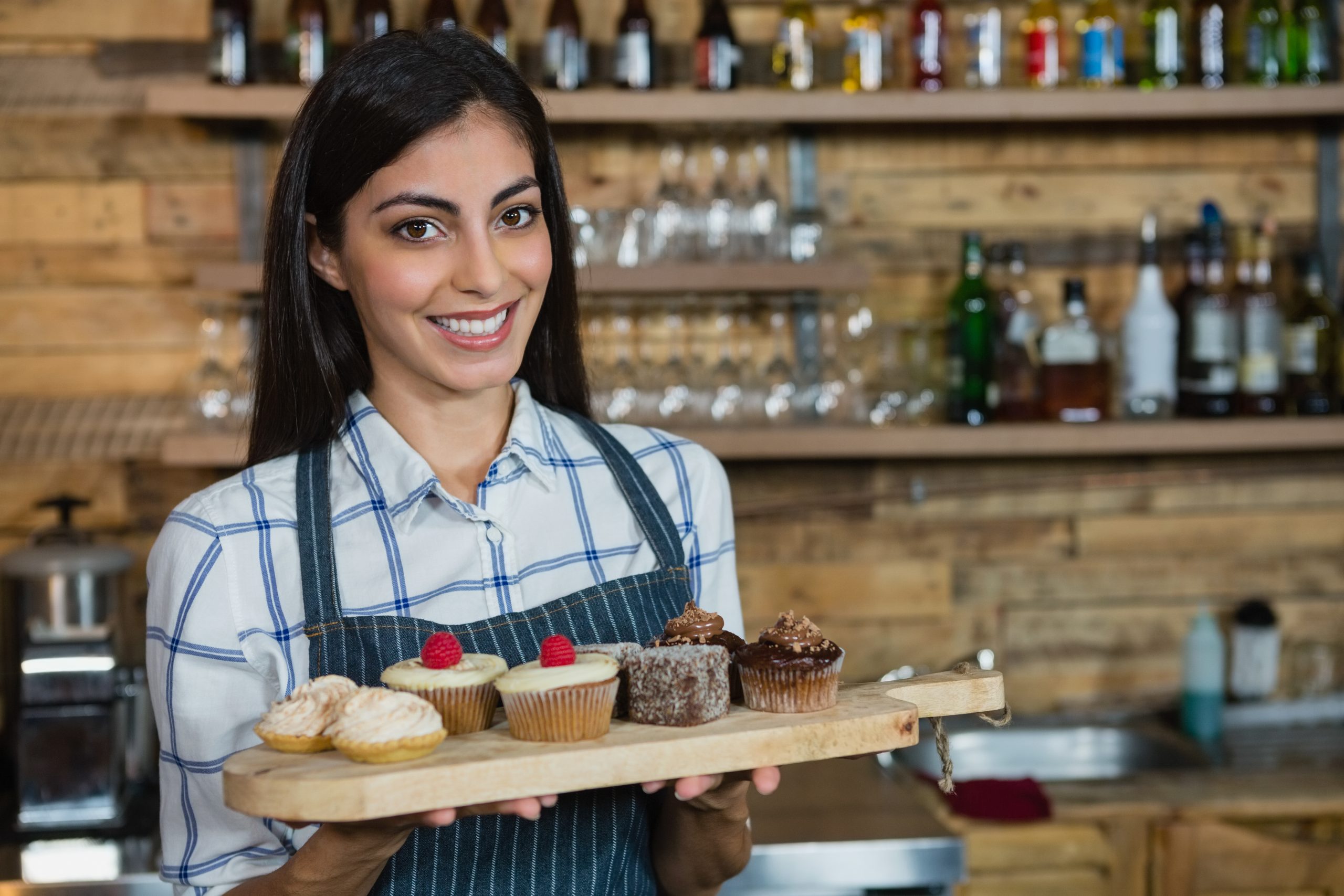 Portrait of smiling waitress holding cupcakes in wooden tray at counter in cafÃ©